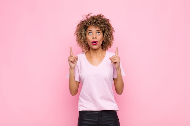 Photo jeune femme afro-américaine se sentir impressionné et bouche ouverte pointant vers le haut avec un regard choqué et surpris sur le mur rose