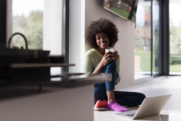 Jeune femme afro-américaine souriante assise sur le sol près d'une fenêtre lumineuse tout en regardant un ordinateur portable ouvert et tenant une tasse à la maison