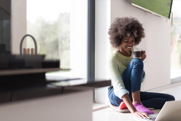 Jeune femme afro-américaine souriante assise sur le sol près d'une fenêtre lumineuse tout en regardant un ordinateur portable ouvert et tenant une tasse à la maison