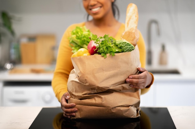 Une jeune femme afro-américaine souriante tient un sac en papier avec des provisions dans un intérieur de cuisine moderne