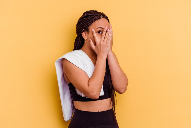 Jeune femme afro-américaine de sport tenant une serviette isolée sur un mur jaune clignote à travers les doigts effrayés et nerveux.