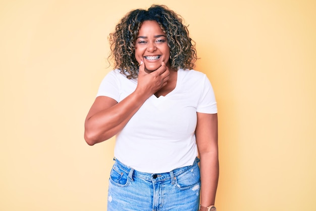 Jeune femme afro-américaine de taille plus portant un t-shirt blanc décontracté regardant confiant à la caméra souriant avec les bras croisés et la main levée sur le menton pensant positif