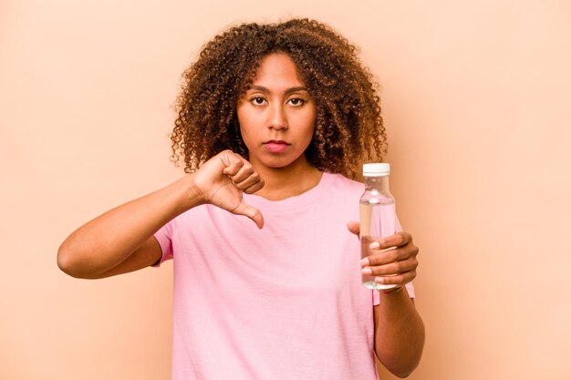Jeune femme afro-américaine tenant une bouteille d'eau isolée sur fond beige montrant un geste d'aversion pouce vers le bas Concept de désaccord