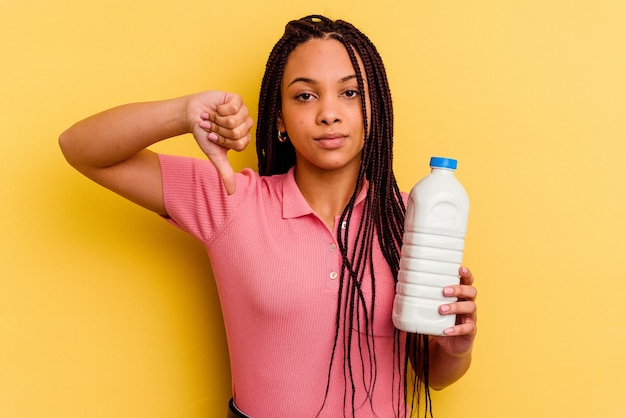 Jeune femme afro-américaine tenant une bouteille de lait isolée sur jaune montrant un geste d'aversion, les pouces vers le bas. Concept de désaccord.