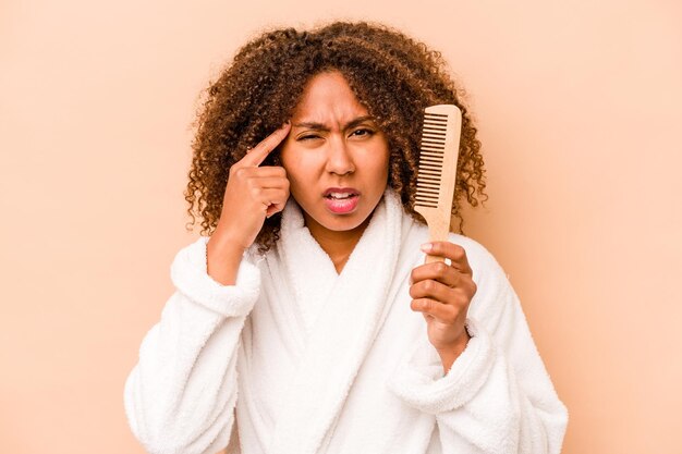 Jeune femme afro-américaine tenant une brosse à cheveux isolée sur fond beige montrant un geste de déception avec l'index