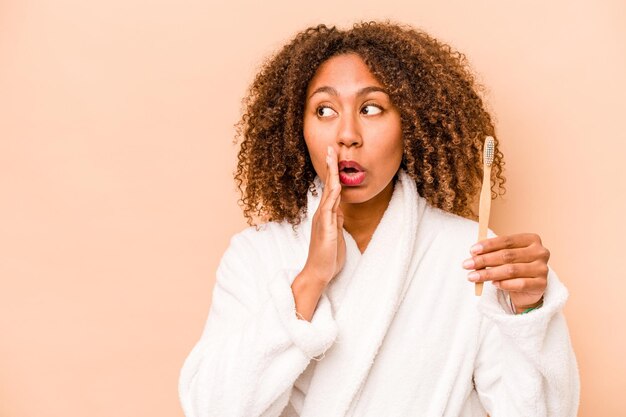 Une jeune femme afro-américaine tenant une brosse à dents isolée sur fond beige dit une nouvelle secrète sur le freinage à chaud et regarde de côté