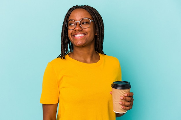 Jeune femme afro-américaine tenant un café à emporter isolé sur fond bleu regarde de côté souriante, gaie et agréable.