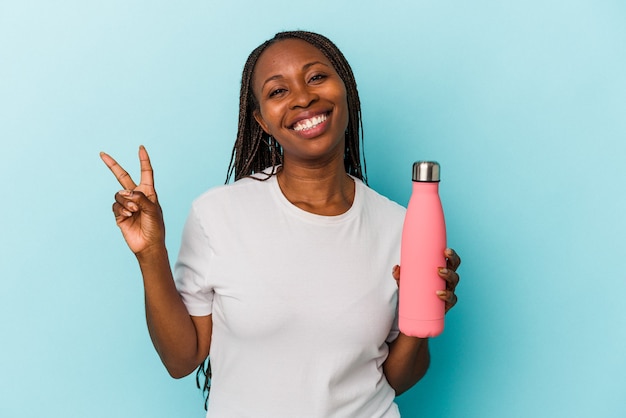 Jeune femme afro-américaine tenant une cantine isolée sur fond bleu joyeuse et insouciante montrant un symbole de paix avec les doigts.