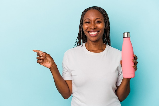 Jeune femme afro-américaine tenant une cantine isolée sur fond bleu souriant et pointant de côté, montrant quelque chose dans un espace vide.