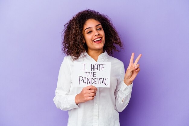 Jeune femme afro-américaine tenant je déteste la pancarte de pandémie isolée sur le mur violet joyeux et insouciant montrant un symbole de paix avec les doigts.