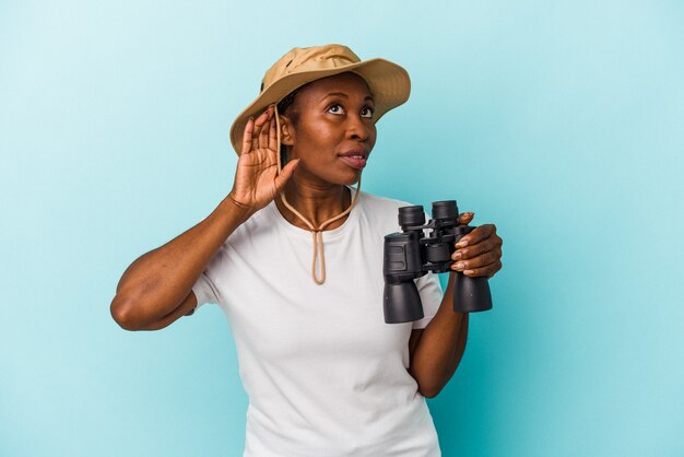 Jeune femme afro-américaine tenant des jumelles isolées sur fond bleu essayant d'écouter un potin.