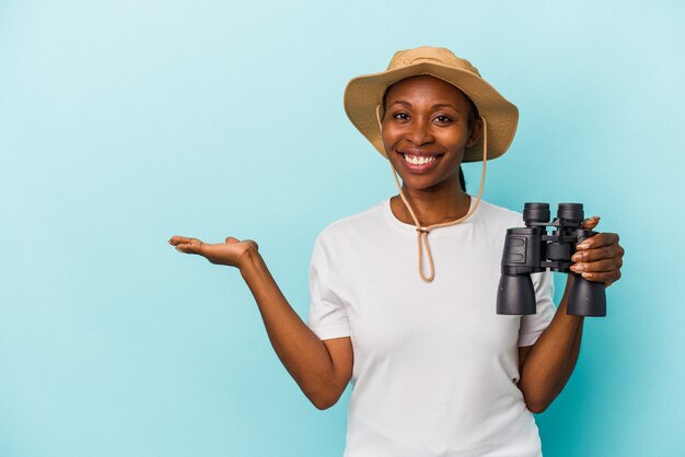 Jeune femme afro-américaine tenant des jumelles isolées sur fond bleu montrant un espace de copie sur une paume et tenant une autre main sur la taille.