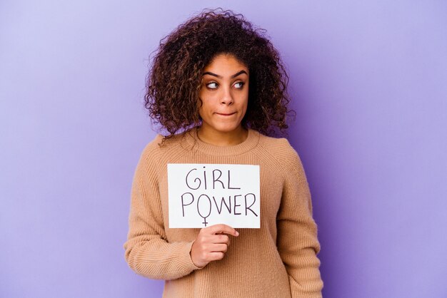 Jeune femme afro-américaine tenant une pancarte Girl power isolée sur un mur violet confuse, se sent douteuse et incertaine.