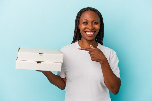 Jeune femme afro-américaine tenant des pizzas isolées sur fond bleu souriant et pointant de côté, montrant quelque chose dans un espace vide.