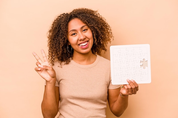 Photo jeune femme afro-américaine tenant un puzzle isolé sur fond beige joyeux et insouciant montrant un symbole de paix avec les doigts