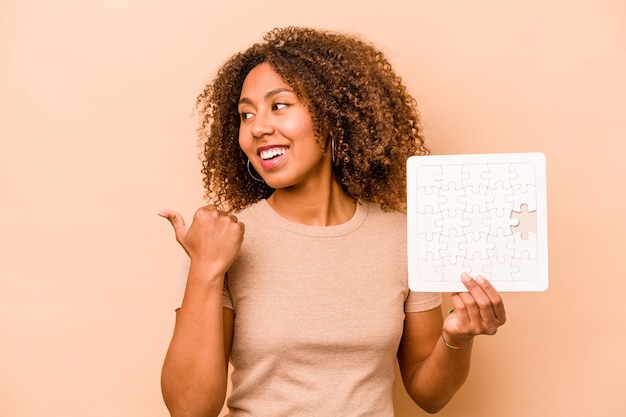 Jeune femme afro-américaine tenant un puzzle isolé sur fond beige pointe avec le pouce en riant et sans souci