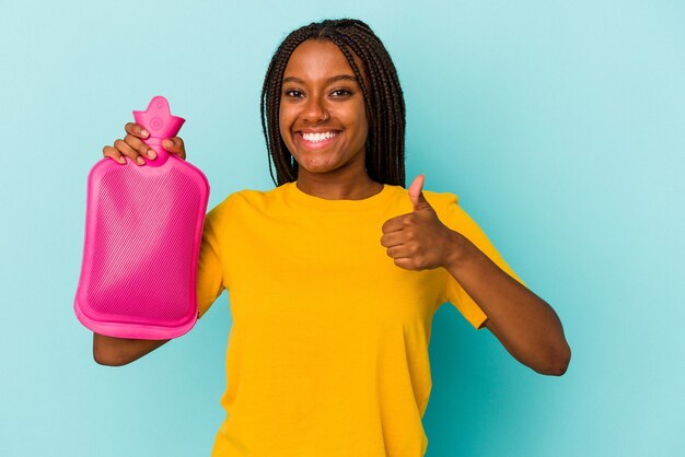 Jeune femme afro-américaine tenant un sac chaud d'eau isolé sur fond bleu souriant et levant le pouce vers le haut