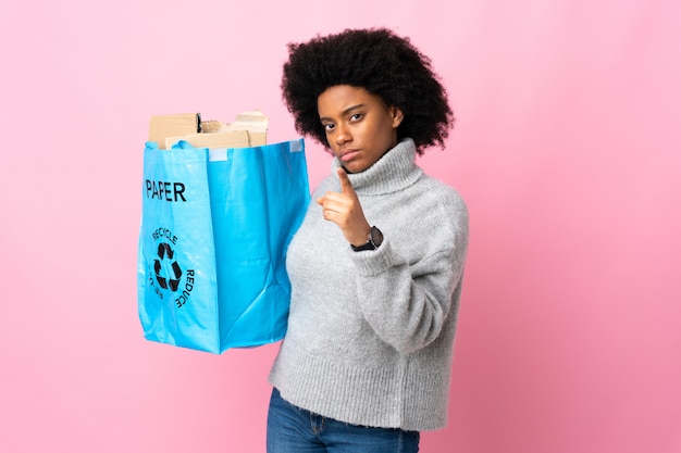 Jeune femme afro-américaine tenant un sac de recyclage isolé sur coloré frustré et pointant vers l'avant