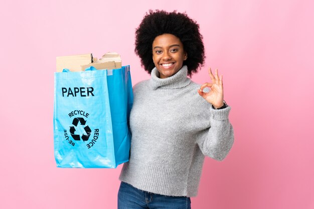 Jeune femme afro-américaine tenant un sac de recyclage isolé sur mur coloré montrant signe ok avec deux mains