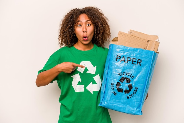 Jeune femme afro-américaine tenant un sac de recyclage plein de papier à recycler isolé sur fond blanc pointant vers le côté