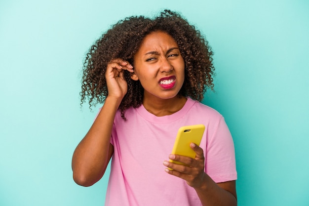 Jeune femme afro-américaine tenant un téléphone portable isolé sur fond bleu couvrant les oreilles avec les mains.