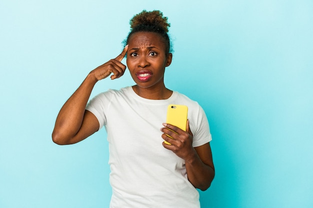 Jeune femme afro-américaine tenant un téléphone portable isolé sur fond bleu montrant un geste de déception avec l'index.