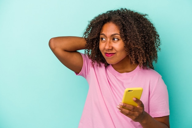 Jeune femme afro-américaine tenant un téléphone portable isolé sur fond bleu touchant l'arrière de la tête, pensant et faisant un choix.