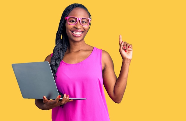 Jeune femme afro-américaine travaillant à l'aide d'un ordinateur portable surpris par une idée ou une question pointant du doigt avec un visage heureux numéro un