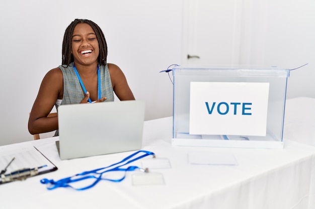 Photo jeune femme afro-américaine travaillant à l'élection politique assis par bulletin de vote souriant et riant fort parce que drôle de blague folle avec les mains sur le corps