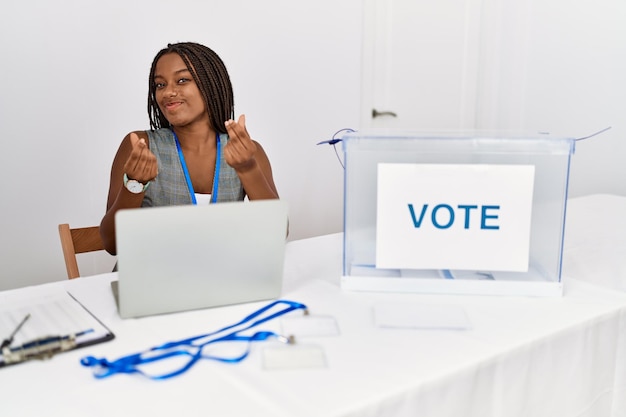 Jeune femme afro-américaine travaillant à une élection politique assise par bulletin de vote faisant un geste d'argent avec les mains, demandant le paiement d'un salaire, une entreprise millionnaire