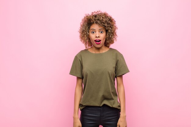 Jeune femme afro-américaine à très choqué ou surpris, regardant avec la bouche ouverte en disant wow contre le mur rose