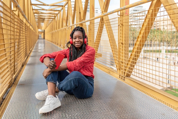 Une jeune femme afro-américaine en tresses assise sur un pont jaune écoutant de la musique avec ses écouteurs rouges et son téléphone portable regardant la caméra