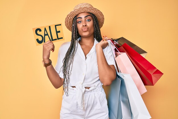 Jeune femme afro-américaine avec des tresses tenant des sacs à provisions et une bannière de vente en regardant la caméra soufflant un baiser étant belle et sexy. expression d'amour.