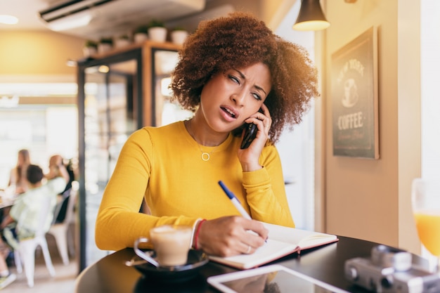 Jeune Femme Afro Au Café, Parlant Au Téléphone Mobile, Très Décontenancée Et Dépassée, écrivant