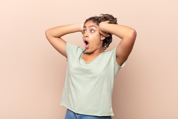 Jeune femme afro avec la bouche ouverte, l'air horrifié et choqué à cause d'une terrible erreur, levant les mains à la tête