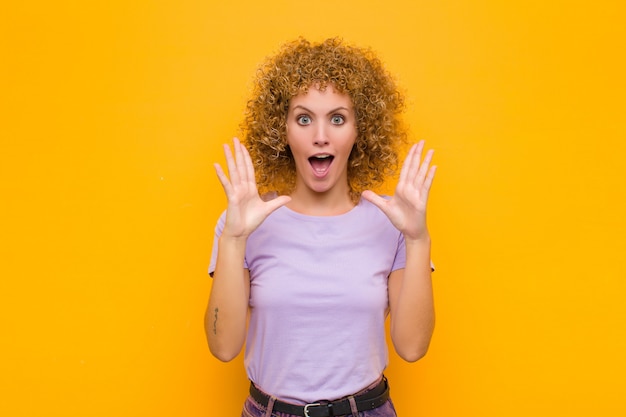 Jeune femme afro, choquée et étonnée, avec la mâchoire tombée de surprise en réalisant quelque chose d'incroyable sur le mur orange