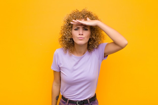 Jeune femme afro à la déroute et étonné, avec la main sur le front à la recherche au loin, regarder ou chercher sur le mur orange