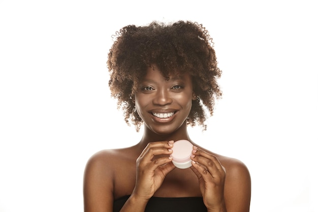 Jeune femme afro-ethnique joyeuse avec une peau propre et saine tenant un pot de crème hydratante