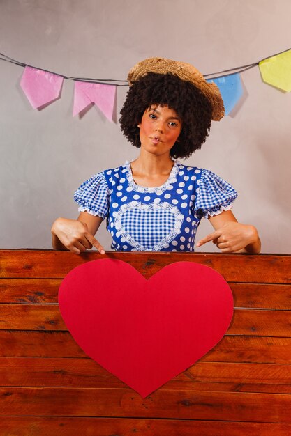 Jeune femme afro habillée pour la fête junina derrière une planche de bois avec un coeur