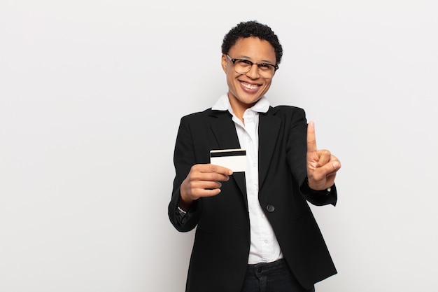 Jeune femme afro noire souriant fièrement et en toute confiance faisant le numéro un pose triomphalement, se sentant comme un leader