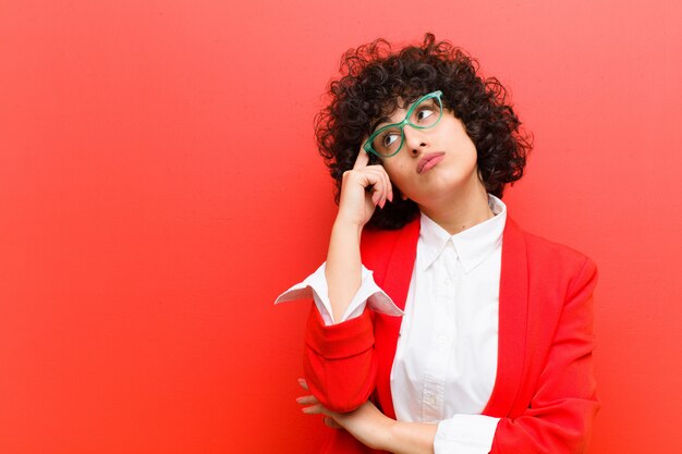 Photo jeune femme afro avec un regard concentré, se demandant avec une expression douteuse, levant les yeux et à côté