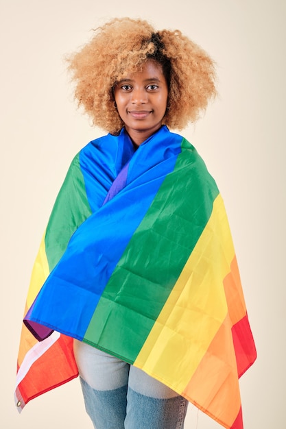 Photo jeune femme afro regardant dans la caméra tout en couvrant d'un drapeau de la fierté gaie sur un fond isolé. concept de communauté lgbtq.