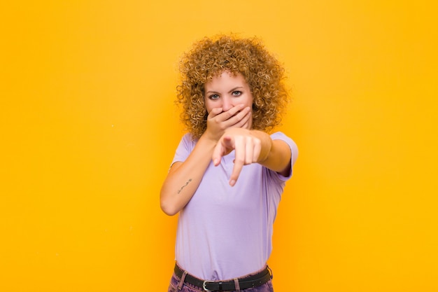 Jeune femme afro se moquant de vous, pointant vers la caméra et se moquant de vous ou se moquant de vous contre le mur orange