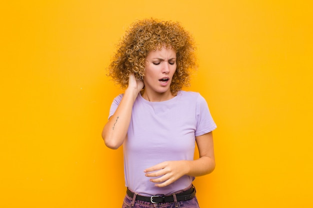 Jeune femme afro se sentant stressée, frustrée et fatiguée, se frottant le cou douloureux, avec un regard inquiet et troublé contre le mur orange