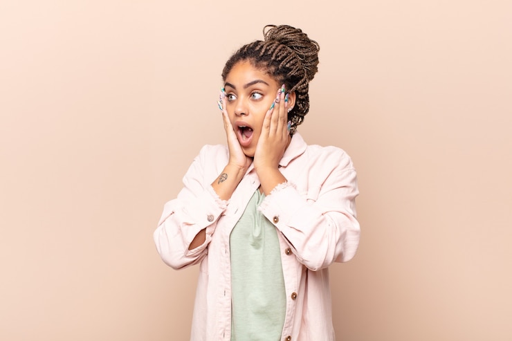 Photo jeune femme afro se sentir heureuse, excitée et surprise, regardant sur le côté avec les deux mains sur le visage
