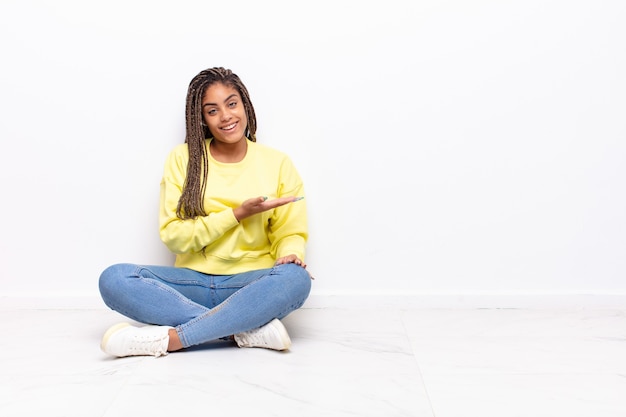 Jeune femme afro souriant joyeusement, se sentir heureux et montrant un concept dans l'espace de copie avec la paume de la main