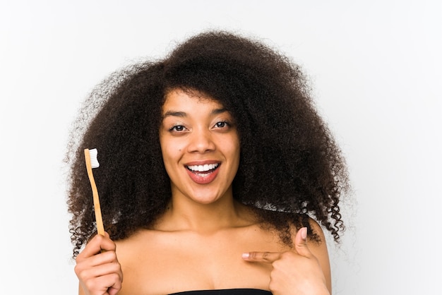 Jeune femme afro tenant une brosse à dents isolée surprise pointant vers lui-même, souriant largement.