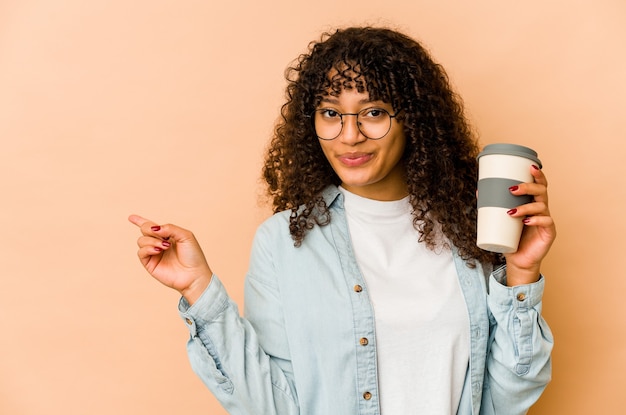 Jeune femme afro tenant un café à emporter souriant et pointant de côté, montrant quelque chose à l'espace vide