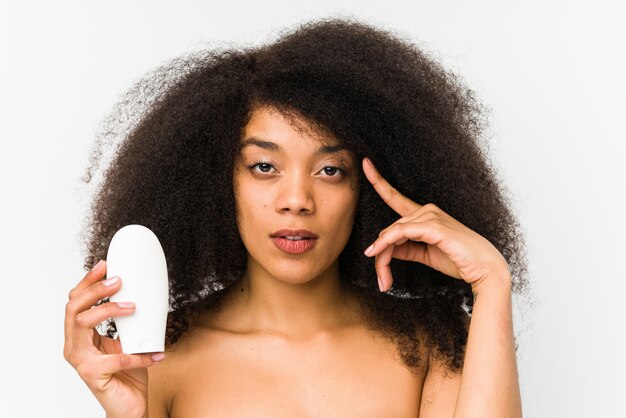 Jeune femme afro tenant une crème hydratante isolée pointant sa tempe avec le doigt, pensant, concentré sur une tâche.