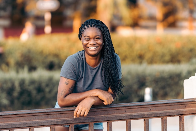 Jeune femme afro avec des vêtements d'été s'appuyant sur un rail tout en souriant à la caméra à l'extérieur